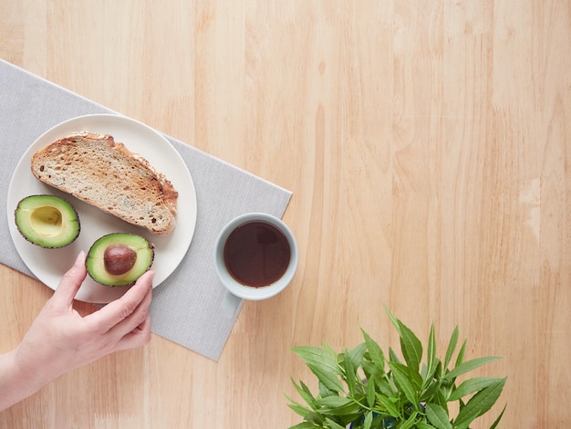 Main de femme tenant un demi-avocat sur une assiette avec du pain grillé à la table du petit-déjeuner Vue de dessus avec espace de copie