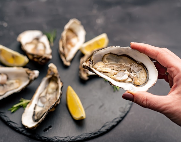 Main de femme tenant une huître fraîchement ouverte tirée d'une assiette noire avec des fruits de mer et du citron