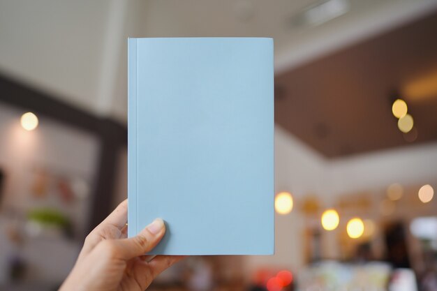 Main de femme tenant un livre bleu clair avec couverture vierge pour insérer du texte sur fond de café flou