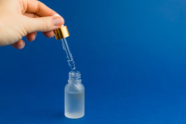 Main de femme tenant une pipette avec de l'huile. Flacon cosmétique avec pipette sur fond bleu tendance. Concept de science ou de test médical. Couleur de l'année 2020. Maquette de bouteille en verre compte-gouttes.