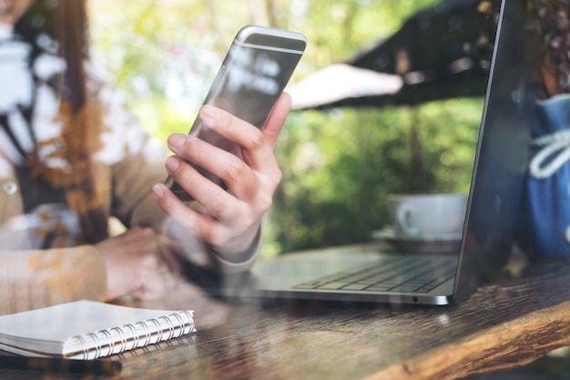 main de femme tenant et regardant un téléphone intelligent tout en utilisant un ordinateur portable au café