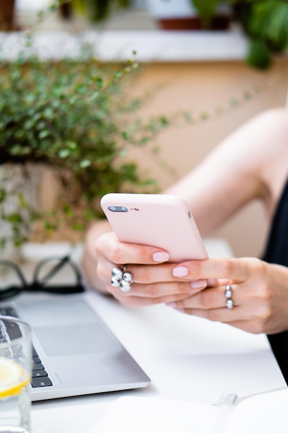 Main de femme tenant un smartphone dans un bureau d'été confortable avec un ordinateur portable et des plantes vertes