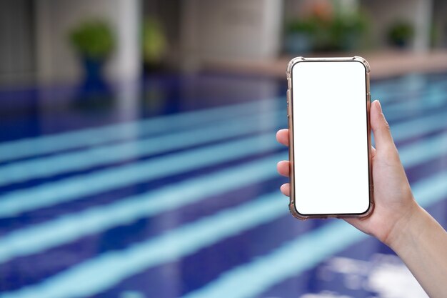 Main de femme tenant un téléphone mobile avec une maquette d'écran blanc vierge au bord de la piscine.