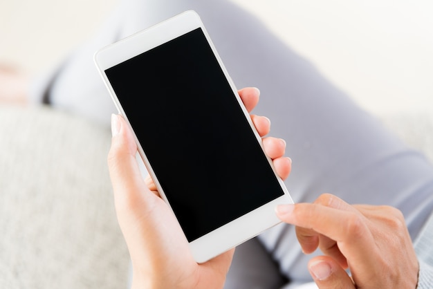 Main de femme tenant un téléphone portable blanc et assis sur un canapé à la maison