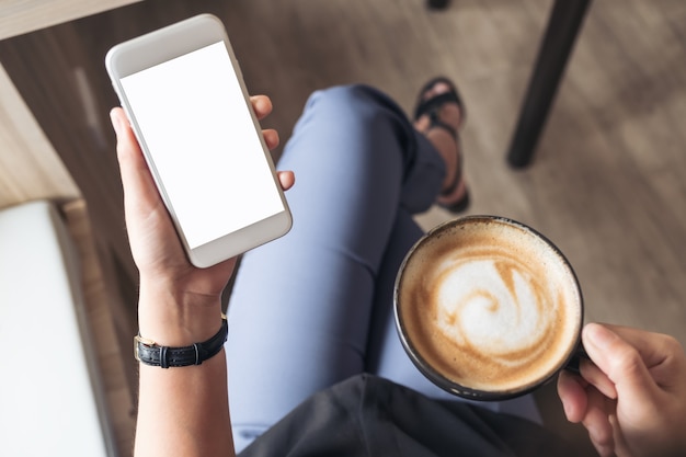 Main de femme tenant un téléphone portable blanc avec écran de bureau vide tout en buvant du café