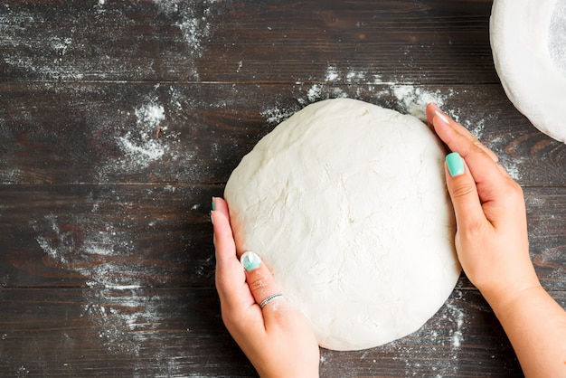 Main de femme tenir la pâte fraîche et le grattoir pour préparer du pain fait maison sur du bois sombre.