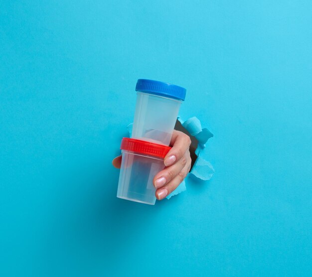 La main de la femme tient un pot en plastique vide pour des tests d'urine. La main sort d'un trou déchiré sur un fond bleu.