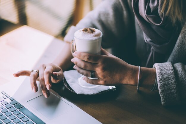 La main d'une femme tient une tasse de café et appuie sur les touches de l'ordinateur portable avec l'autre main