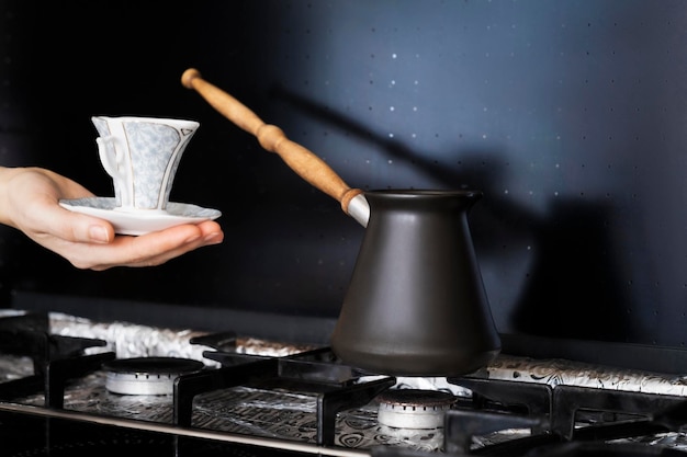 la main d'une femme tient une tasse de café avec une soucoupe près d'une cuisinière à gaz sur laquelle le café est infusé dans une cera