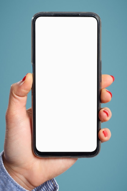 La main d'une femme tient verticalement un smartphone avec un écran blanc vierge sur fond bleu. Pour le copyspace.