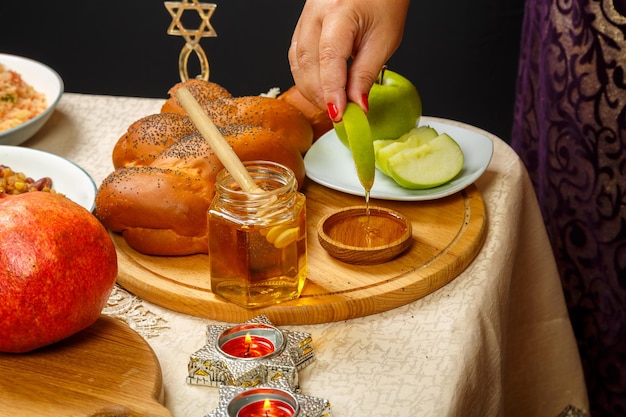 La main d'une femme trempe un morceau de pomme dans du miel en l'honneur de la célébration de Rosh Hashanah près du miel et de la challah et de la nourriture traditionnelle