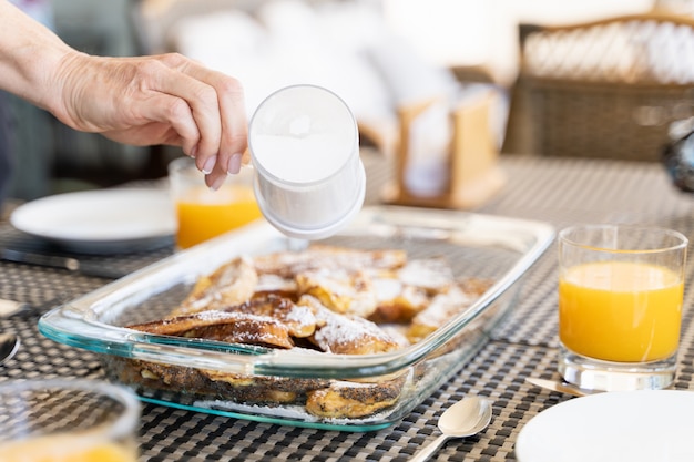 Main de femme versant du sucre en poudre sur un plateau en verre de pain perdu par des jus d'orange sur la table du petit-déjeuner