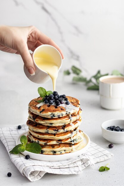 La main de la femme verse du lait condensé sur une pile de crêpes avec des bleuets frais sur du marbre