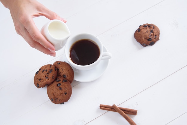 La Main D'une Femme Verse Du Lait Dans Le Café. Une Tasse De Café Parfumé à La Cannelle Et Biscuits Sur Fond Blanc. Vue De Dessus.