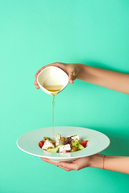 Une main de femme verse de l'huile d'olive dans une salade de feta grecque sur fond turquoise