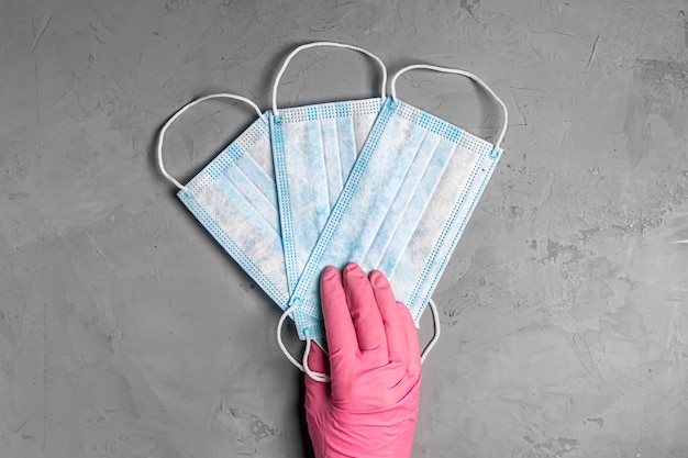 La main des femmes dans des gants roses contient trois masques médicaux jetables bleus sur fond gris. Mains de femme avec gros plan de masque. Vue de dessus, mise à plat.