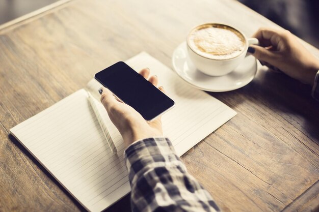 Main de fille avec journal vierge de smartphone et tasse de café sur une table en bois