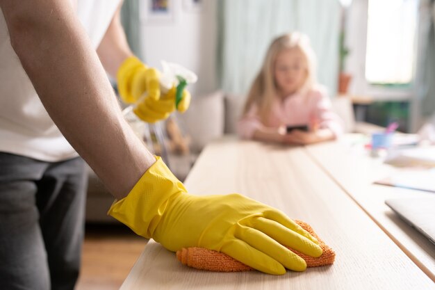 Main gantée de jeune homme frottant la table en bois avec un chiffon tout en le nettoyant avec un détergent avec sa petite fille assise sur l'arrière-plan