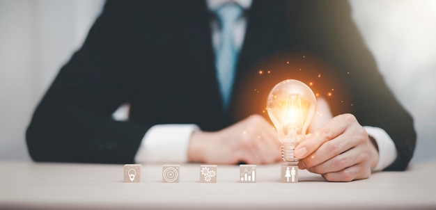 Main d'homme d'affaires tenant une ampoule avec icône sur un cube en bois et travaillant sur un bureau La créativité et l'innovation sont les clés du succèsConcept de nouvelle idée et d'innovation avec le cerveau et les ampoules fonctionnant a