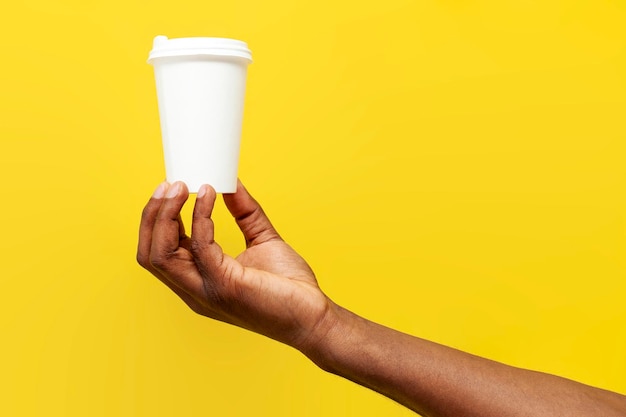 La main d'un homme afro-américain tient une tasse de café en papier blanc sur un fond jaune.