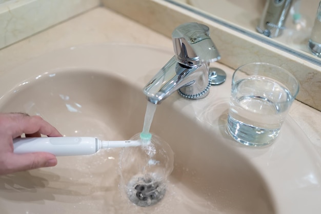 La main de l'homme blanc lave la brosse à dents électrique dans le lavabo de la salle de bain