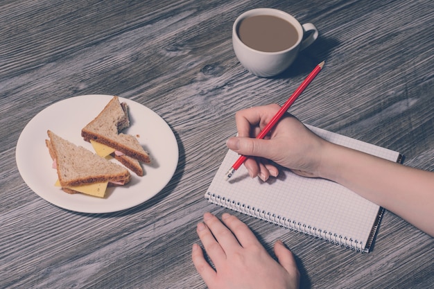 La main de l'homme effaçant quelque chose dans un cahier. Fond de savoureux sandwich au fromage mordu et tasse de café chaud. Gros plan, style vintage