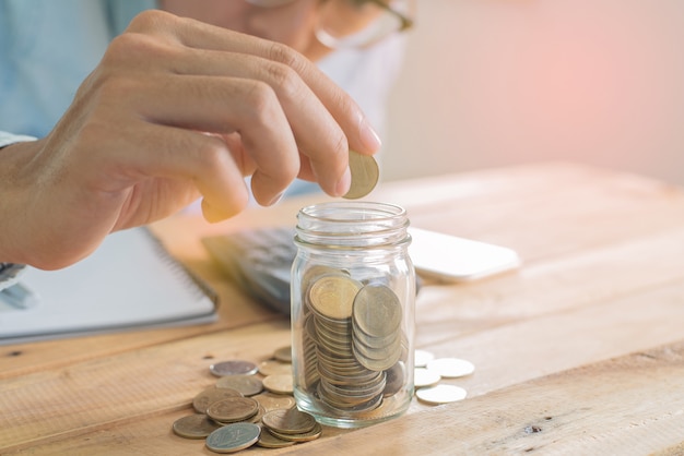 La main de l'homme mettant la pièce d'économie en verre sur une table de travail en bois avec un tas de pièces de monnaie, un ordinateur portable, un smartphone et un graphique - Investissement, affaires, finance et banque