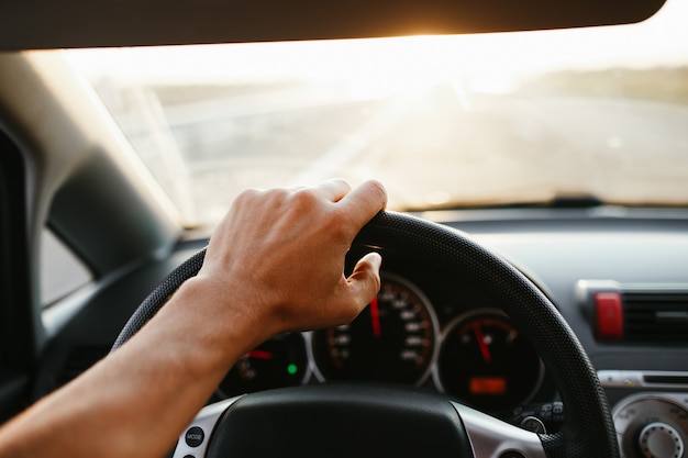 La main de l'homme de mise au point sélective sur le volant, conduisant une voiture au coucher du soleil. Fond de voyage.