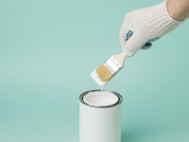 La main d'un homme avec un pinceau avec de la peinture blanche sur un pot de peinture sur fond bleu. Exécution des travaux de peinture.