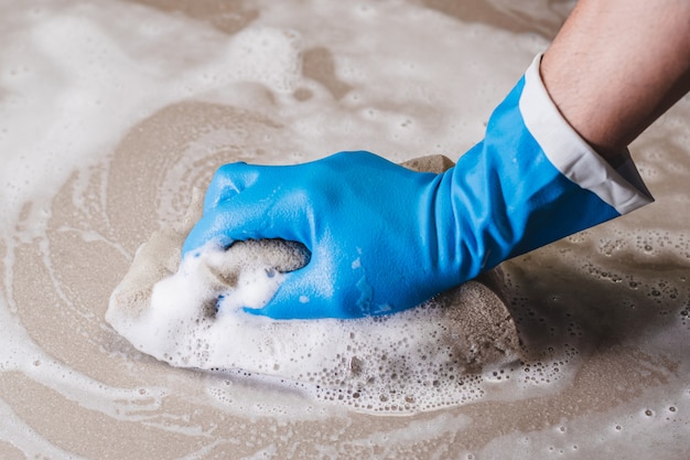 Main d'homme portant des gants en caoutchouc bleu utilise une éponge pour nettoyer le carrelage.