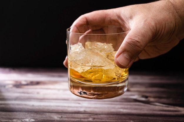 La main d'un homme soulevant un verre de whisky ou de scotch et de gros morceaux de glace d'une table en bois rustique