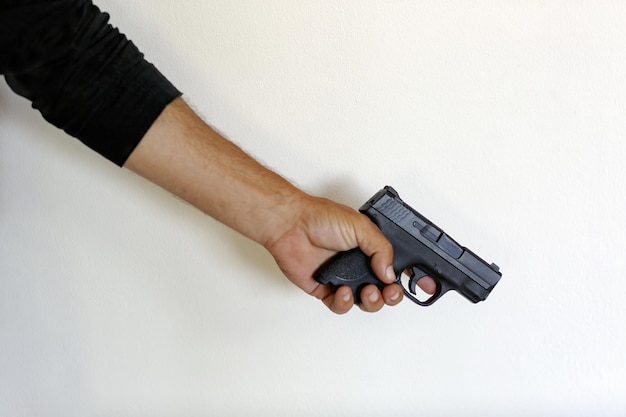Photo main d'homme tenant une arme sur un fond mural blanc main d'homme tenant une arme de poing 9 mm tueur criminel illégal close up de l'homme main avec pistolet noir doigt sur la gâchette de l'arme focalisation sélective