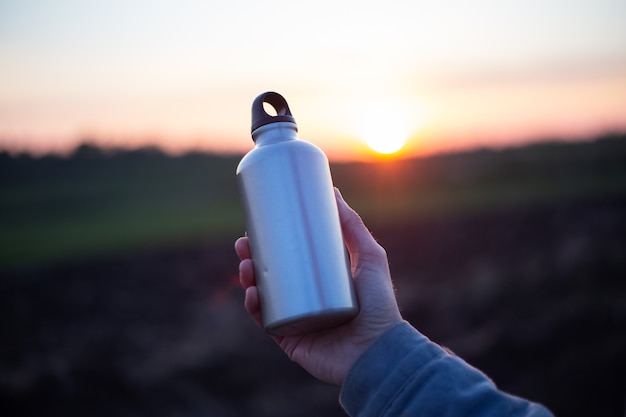 Main d'homme tenant une bouteille en aluminium pour l'eau, sur fond de coucher de soleil