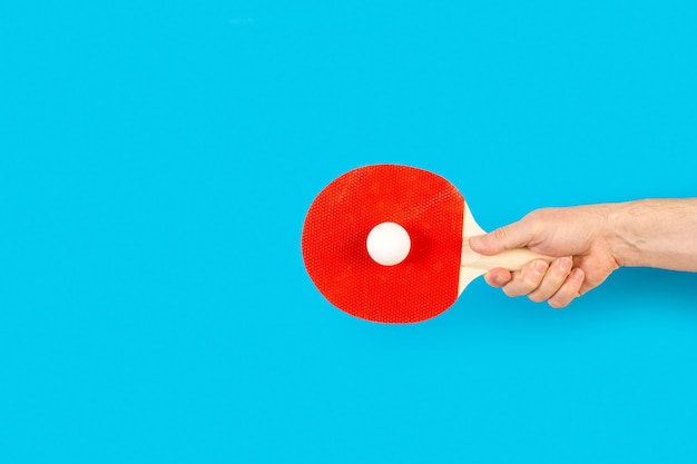 Main d'homme tenant une raquette de ping-pong rouge avec une balle blanche sur fond bleu