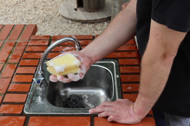 La main d'un homme tient une éponge savonneuse sur un évier dans une cuisine en plein air