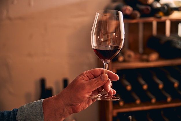 main de l'homme avec un verre de boisson alcoolisée et des casiers à vin sur fond flou