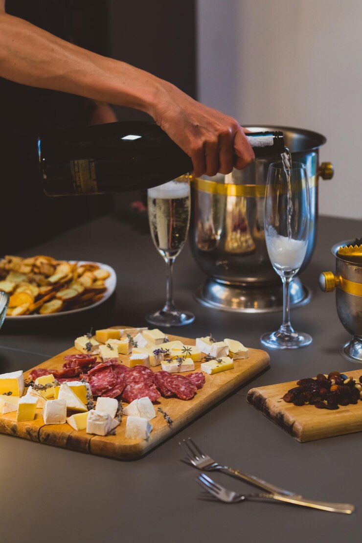 Photo la main de l'homme versant du champagne dans une planche de bois en verre avec du camembert coupé et du salami ensemble de collations simples et savoureuses sur des planches en bois deux verres avec seau à champagne pour refroidir les boissons