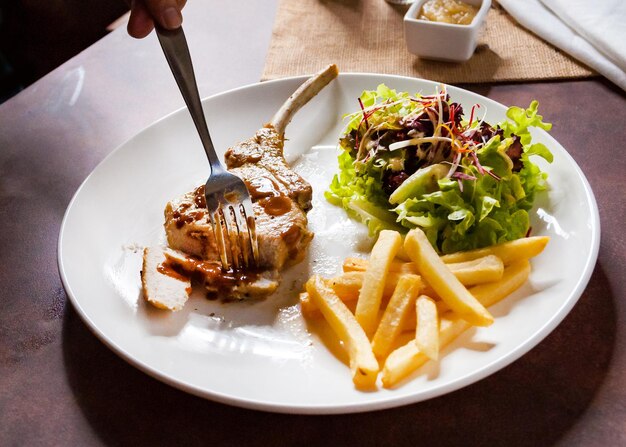 Main humaine utilisant un couteau coupant un steak de côtel de porc grillé de près dans un restaurant Steak de coupe de porc avec salade et frites