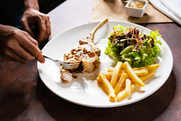 Main humaine utilisant un couteau coupant un steak de côtel de porc grillé de près dans un restaurant Steak de coupe de porc avec salade et frites