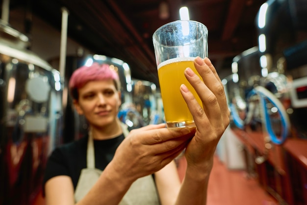 Main de jeune brasseuse en tablier examinant un verre de bière fraîche après préparation en se tenant debout parmi d'énormes réservoirs devant la caméra