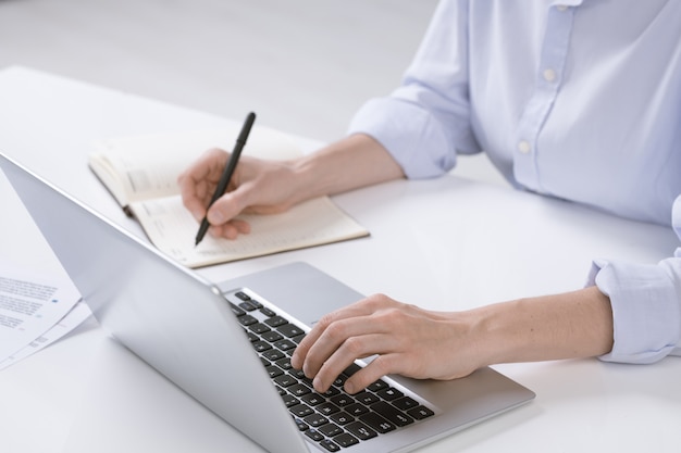 Main de jeune femme d'affaires en poussant les touches du clavier d'ordinateur portable tout en prenant des notes dans l'ordinateur portable par lieu de travail