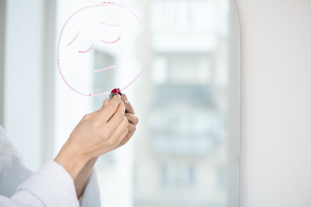 Main de jeune femme en peignoir avec rouge à lèvres dessin visage bouleversé sur miroir tout en exprimant son désespoir