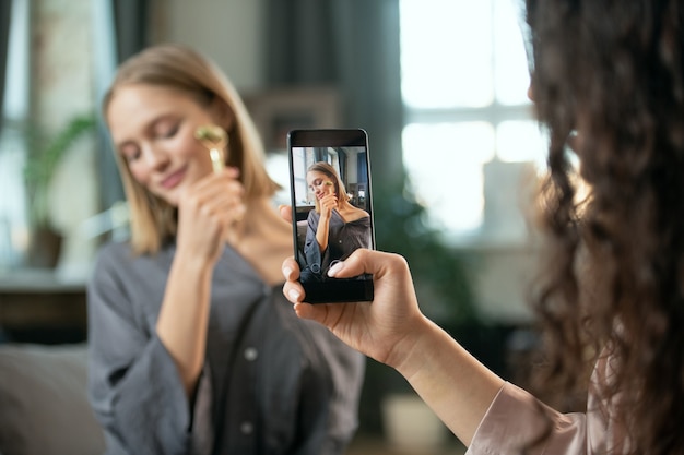 Main de jeune femme avec un smartphone tirant sur son amie à l'aide d'un rouleau de massage sur son visage et profitant d'une procédure de beauté