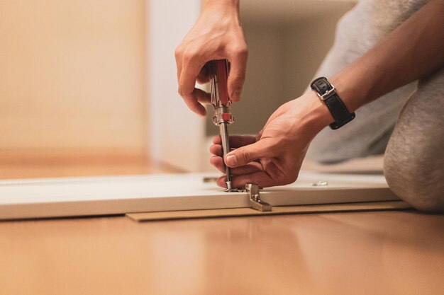 Main de jeune homme serrant une vis avec un tournevis sur une porte d'armoire en bois blanc