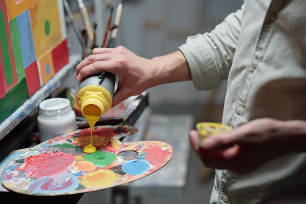 Main de jeune peintre ajoutant de la couleur jaune sur la palette avant de commencer à peindre en se tenant debout devant le chevalet en studio d'arts