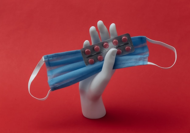 Une main de mannequin blanche tient un masque médical et un blister de pilules sur fond rouge. Concept de minimalisme de médecine