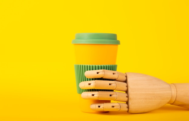 Main de mannequin en bois tenant une tasse de café réutilisable sur une surface jaune