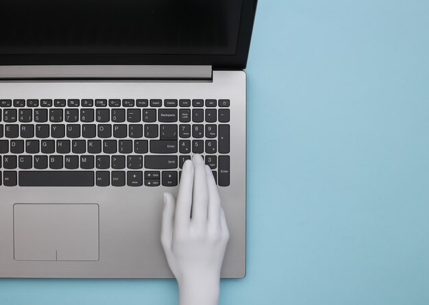 Main de mannequin tapant sur le clavier d'un ordinateur portable. Fond bleu. Vue de dessus, minimalisme