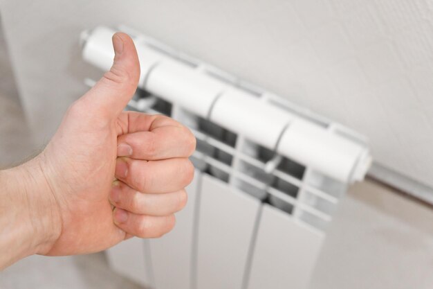 La main masculine montre le signe du pouce vers le haut contre le radiateur de chauffage domestique installé