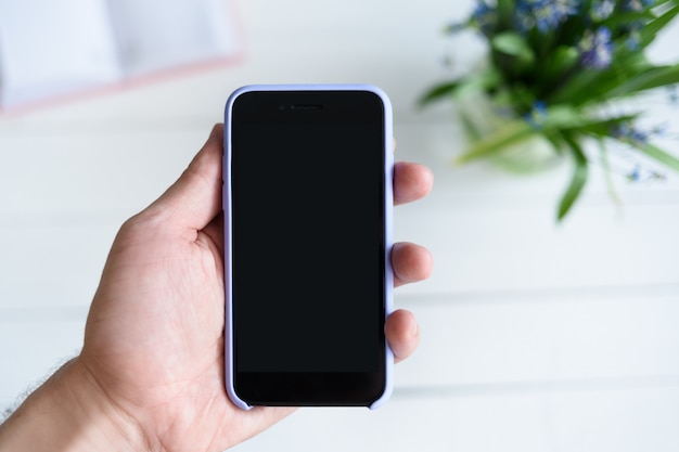 Main masculine avec un smartphone. Écran vide noir. Table avec carnet et fleurs